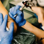 mani con guanto monouso celesti che tatuano un braccio