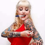 donna mezzo busto bionda vestita di rosso conun pennarello in bocca e uno in mano col quale si colora le braccia comp0letamente tatuate